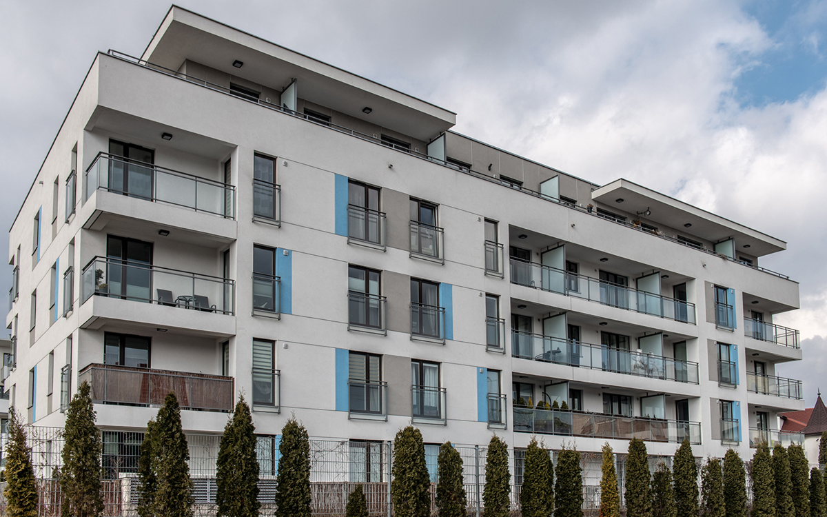 Zdjęcie. Elewacja fasady jednego z budynków osiedla Wille Lazurowa. Zbudowana z pomocą płyt cementowych. Balkony obudowane przezroczystą balustradą. Budynek jest biały, z niebieskimi prostokątami przy prostokątnych oknach.
