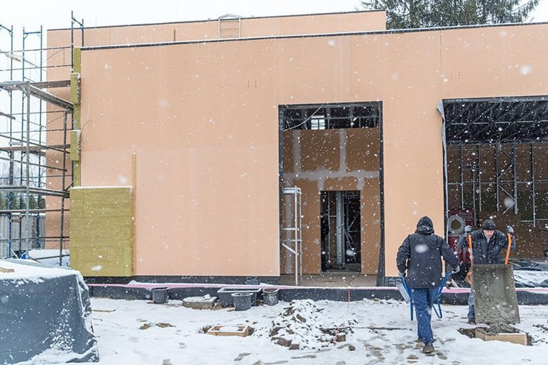 Zdjęcie przedstawiające budynek edukacyjny świetlicy w Nowej Wsi. Budynek zbudowany w technologii szkieletowej z wykorzystaniem konstrukcji stalowej. Widać prostokątną część budynku w kolorze beżowym. Przed budynkiem stoją robotnicy. Pada śnieg.