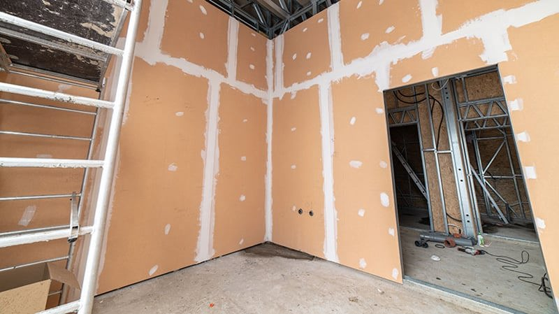 Widok pokoju w czasie budowy w budynku. Po prawej stornie otwór do przejścia do kolejnych pomieszczeń, przez który widać metalowe ożebrowania na ścianach w innych pomieszczeniach. Po lewej stronie kadru białe rusztowanie. Ściany w pokoju są blado pomarańczowe.