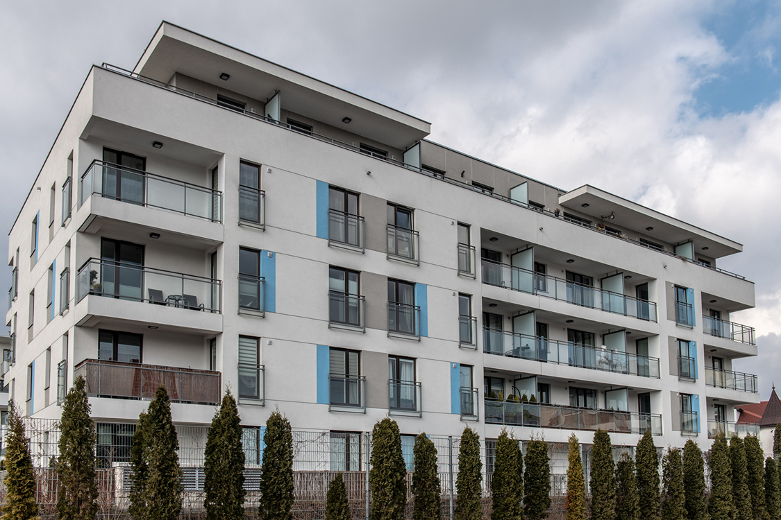 Zdjęcie. Elewacja fasady jednego z budynków osiedla Wille Lazurowa. Zbudowana z pomocą płyt cementowych. Balkony obudowane przezroczystą balustradą. Budynek jest biały, z niebieskimi prostokątami przy prostokątnych oknach.